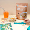 Millet vermicelli package with a glass of juice, salad, and Scrabble game on a table.