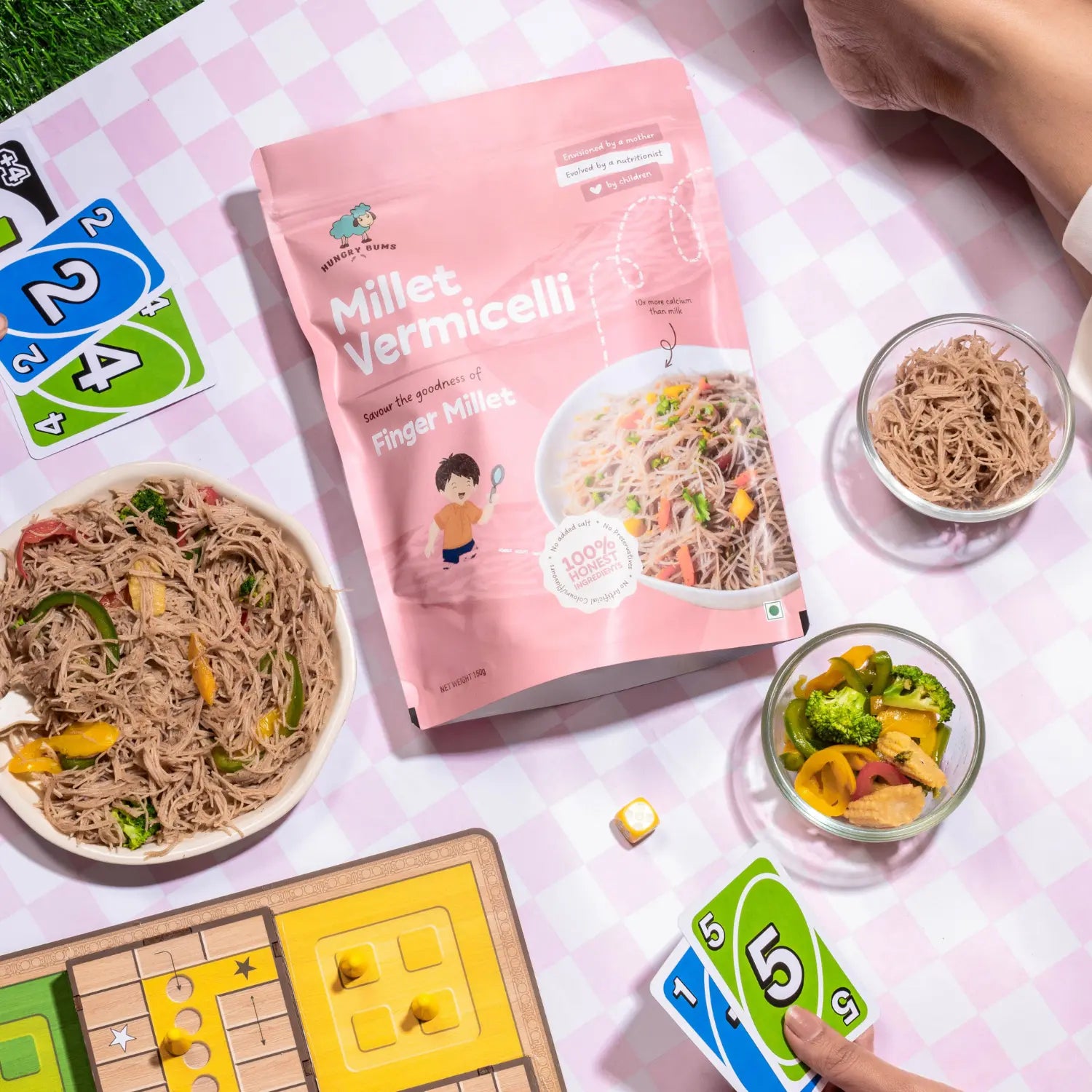 Millet vermicelli package with bowls of noodles and a board game on a checkered tablecloth.