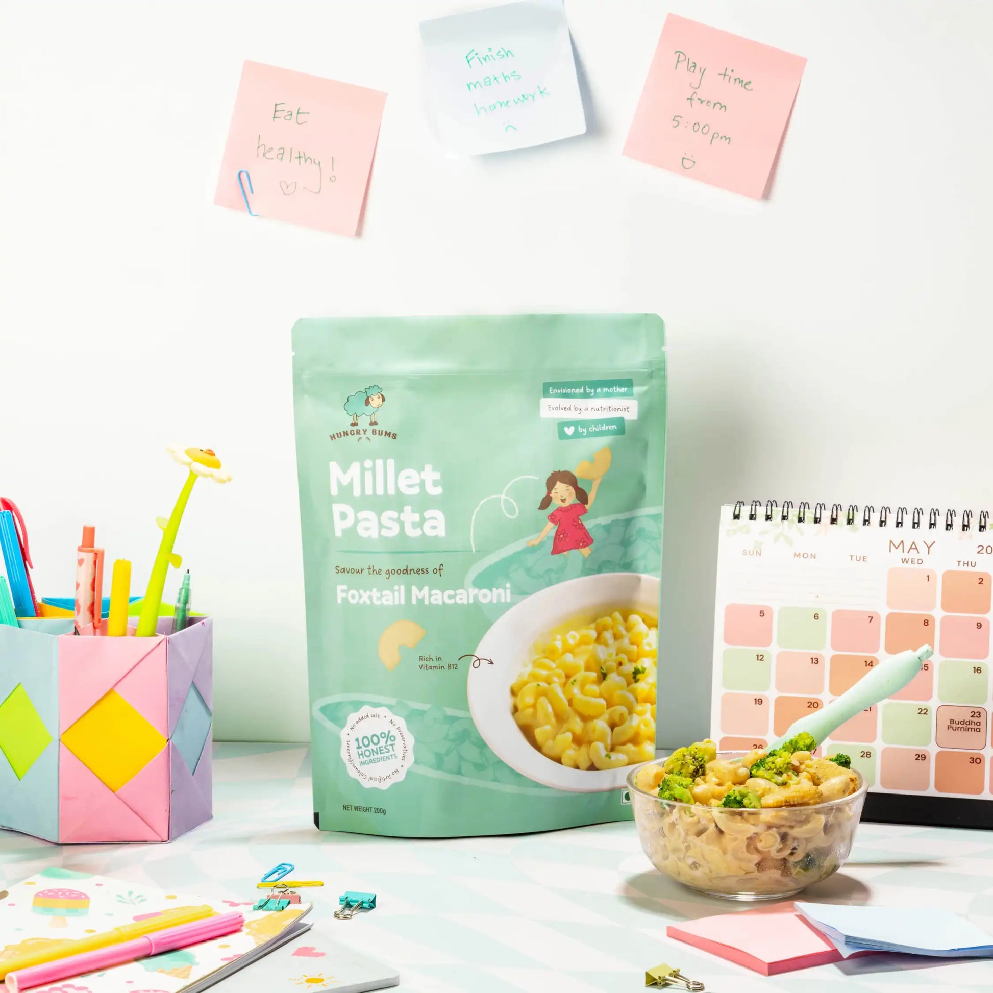 Millet Pasta package on a desk with a bowl of pasta and stationery items.